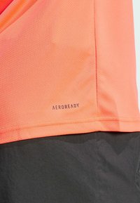 Camisa atlética ligera con textura de malla transpirable, color coral brillante y logo "AEROREADY" en el dobladillo.