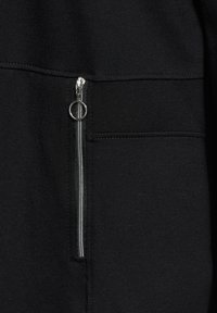 Tessuto nero con una superficie liscia, caratterizzato da una cerniera argento e un anello per tirare tondo sulla tasca. Cuciture visibili lungo le cuciture.