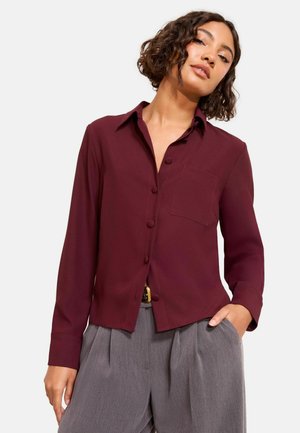 Burgundy button-up shirt with a collar, single chest pocket, long sleeves, and a relaxed fit, paired with gray pleated trousers.