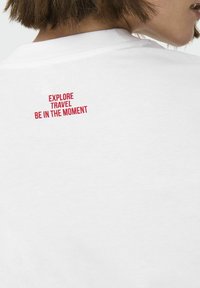 Witte t-shirt met rode tekst op de rug met de tekst "VERKEN REIS BELEEF HET MOMENT," met een eenvoudig ronde halsontwerp en een zachte stoftextuur.