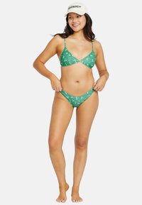 Zelený bikiny set s kvetinovým vzorom, s tenkými nastaviteľnými ramienkami a minimalistickým dizajnom. Model má na sebe svetlú šiltovku.