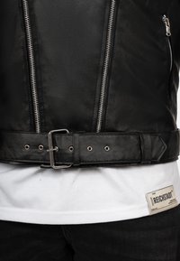 Zwarte leren jas met zilveren ritsen, een taille met riem en metalen gesp, en een gladde textuur. Draagt over een wit T-shirt met een label.