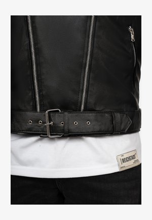 Czarna skórzana kurtka z metalowymi suwakami, z paskiem w talii z metalową klamrą i gładką teksturą. Nosi na sobie białą koszulkę z metką.