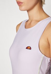 Lilla ermeløs topp laget av glatt stoff. Har et lite brodert logo i rødt, oransje og mørkeblått, med grønne detaljer langs siden.