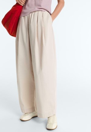 Osoba nosząca luźne beżowe spodnie, kremowe trampki, pasiasty top i trzymająca dużą czerwoną torbę na białym tle.