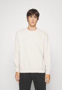Lichtbeige sweatshirt met een gestructureerd ruitpatroon, ronde hals en geribde manchetten. Gekleed met donkere broeken.