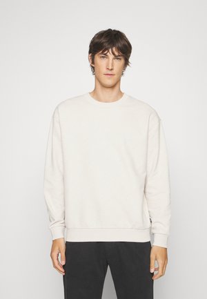 Felpa beige chiaro con un motivo a diamante testurizzato, colletto a giro, e polsini a coste. Abbinata a pantaloni scuri.