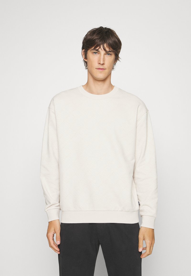 Lichtbeige sweatshirt met een gestructureerd ruitpatroon, ronde hals en geribde manchetten. Gekleed met donkere broeken.
