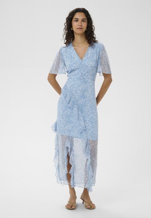 Vrouw met krullend haar, gekleed in een lichtblauwe wikkeljurk met bloemenprint, asymmetrische zoom met ruches en korte vlindermouwen, staand op blote voeten.