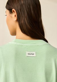 Ljusgrön kortärmad skjorta med rund halsringning och en tygetikett på baksidan med texten "Artist Editions Frankie Thorp." Mjuk textur.