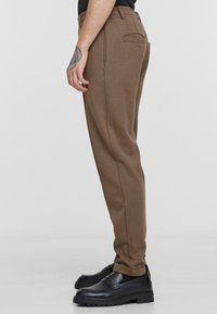 Brown sweatpants with elastic waistband, tapered fit, rear pocket, textured fabric, paired with black loafers featuring a thick sole.
