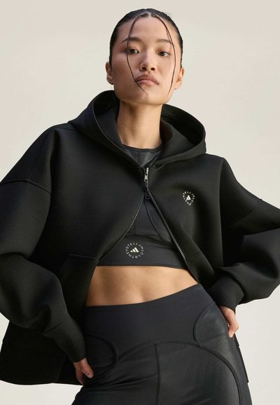 Čierna mikina na zips s krátkym strihom, s logom na hrudi. Nosená nad čiernym športovým topom a legínami s vysokým pásom.