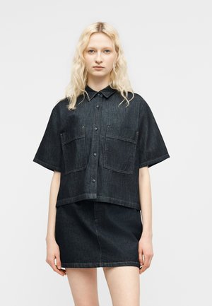 Νεαρή γυναίκα με κυματιστά ξανθά μαλλιά, που φοράει κοντομάνικο πουκάμισο από σκούρο τζιν με κουμπιά και τζιν μίνι φούστα στο ίδιο χρώμα, στέκεται μπροστά σε απλό φόντο.