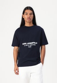 Tengerészkék, rövid ujjú póló fehér nyomtatott szöveggel. Normál fazon, környakú. Pamut anyag, sima tapintású. Fehér nadrág.