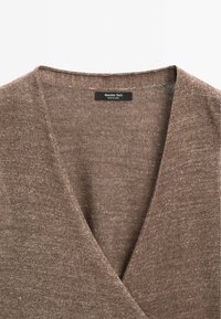 Pull en maille marron avec un col en V. La texture est douce et légèrement chinée, avec une étiquette au niveau de l'encolure. Confectionné en matériaux mélangés.