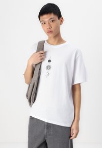 T-shirt bianca in cotone con una grafica che presenta simboli celesti in nero. Il modello indossa una borsa a spalla grigia e pantaloni grigi.
