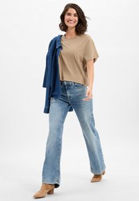 Beige korte mouwen top, lichtblauwe wijde jeans en tan haklaarzen. Een spijkerjack casual over de arm gedrapeerd.