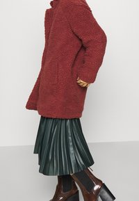 Burgundy fleece-kappa med långa ärmar och lös passform, bärs över en plisserad grön kjol och bruna klumpiga stövlar med klack.