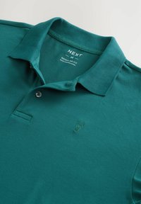 Polo de color verde azulado hecho de tejido texturizado, con un cuello clásico, tapeta de dos botones y un pequeño logotipo bordado en el pecho.