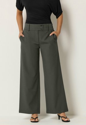 Vrouw draagt donkergrijze, wijde broek met hoge taille en drie knopen, gecombineerd met een zwarte top en zwarte sandalen met hak. Handen in de zakken.