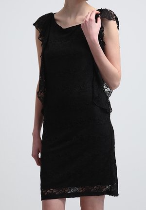 Femme portant une robe noire en dentelle sans manches avec des détails en dentelle volantée le long des côtés et de l'ourlet, debout devant un fond clair uni.