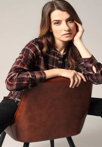 Chaise en simili cuir marron avec un dossier arrondi. Un modèle en chemise à carreaux repose les mains sur la chaise, mettant en valeur le texture et le design de la chaise.