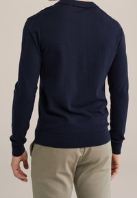 Pulóver de algodón azul marino con mangas largas y dobladillo acanalado, que presenta un cuello clásico y una textura mínima, llevado sobre pantalones de color claro.