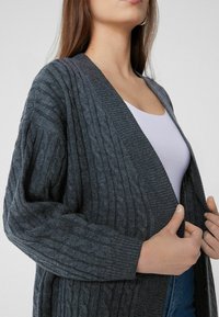 Graue Rippstrickjacke mit lockerer Passform, aus strukturiertem Strickstoff. Offene Vorderseite und weite Ärmel, die eine helllila Innenschicht enthüllen.
