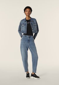 Farkkutakki, jossa on lyhennetty muotoilu, vaaleansininen väri, kaksi rintataskua, mustan paidan päällä, yhdistettynä korkeavyötäröisiin sinisiin farkkuihin ja mustiin lenkkareihin.