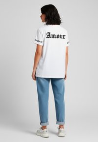 Witte katoenen T-shirt met zwarte tekst "Amour" op de achterkant en "toujours" op de mouwen. Gepaard met lichtblauwe broek met opgerolde mouwen en witte sneakers.