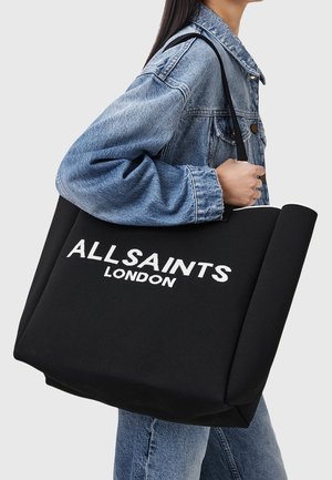 Persoon met een blauwe spijkerjack en spijkerbroek draagt een grote zwarte draagtas met witte tekst "ALLSAINTS LONDON" op de schouder.