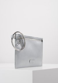 Silverfärgad läderclutch med en slät yta, stort cirkulärt ringaccent, dragkedjestängning och en märkesetikett på framsidan.