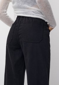 Pantaloni neri a vita alta realizzati in tessuto spesso, con tasche posteriori a toppa e una leggera lucentezza. La vita è elasticizzata per un maggiore comfort.