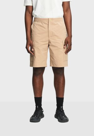Mann trägt beige Cargo-Shorts, weißes T-Shirt, schwarze Socken und schwarze Turnschuhe, steht vor einem schlichten hellen Hintergrund.