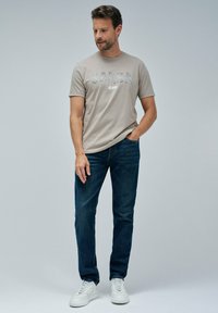 Světle béžové tričko s potiskem "Salsa Jeans" ve třpytivých písmenech, spárované s tmavě modrými džíny a bílými teniskami.