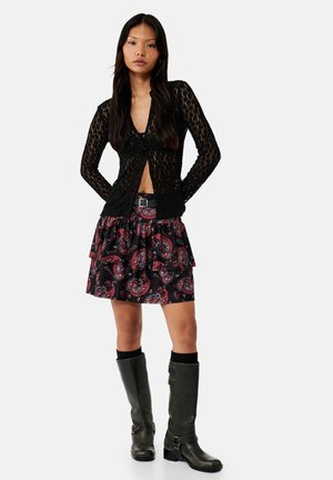 Mujer con una blusa negra de manga larga de encaje, una falda de varias capas con estampado paisley y cinturón, y botas negras hasta la rodilla con hebillas.