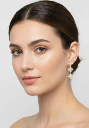 Young woman with smooth skin and dark hair wears elegant dangling pearl and gold floral earrings against a neutral background.