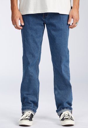 Person wearing blue straight-leg jeans, black and white sneakers, and a white shirt, standing against a plain light background.