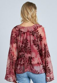 Blusa burgundy transparente con patrón floral, escote con volantes y detalle peplum. Mangas largas con puños de volantes. Llevada con jeans azules.