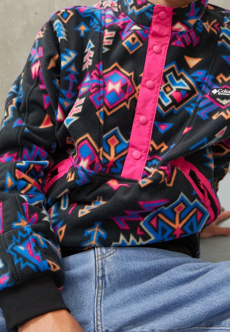 Fleece pullover-takki, jossa on musta perusväri ja värikkäitä geometrisia kuvioita sinisessä, pinkissä ja oranssissa, jotka on korostettu pinkillä nappilistan kanssa.