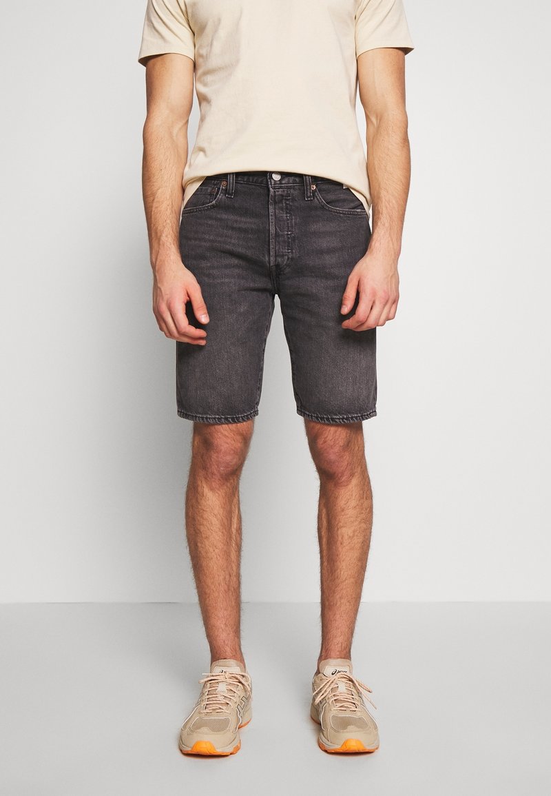 Homme portant un t-shirt beige, un short en jean noir et des baskets beiges avec semelles orange, debout devant un fond uni.