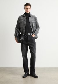 Gray corduroy shirt jacket with two chest pockets, black turtleneck sweater, dark denim jeans, black belt, and black shoes, holding a black bag.