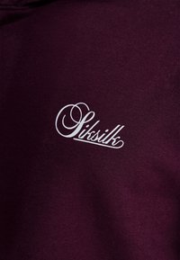 Bordeauxrode sweatshirt van zachte stof, met een wit logo in schrift "Siksilk" op de linkerborst.