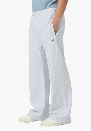 Persona che indossa pantaloni della tuta grigio chiaro larghi con logo Champion sulla coscia, mano in tasca, abbinati a scarpe bianche e maglietta blu.