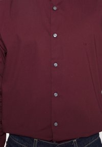 Burgundowa koszula z guzikami, z widocznymi czterema guzikami i haftowanym napisem "CK" w okolicy klatki piersiowej, noszona z ciemnoniebieskimi dżinsami.