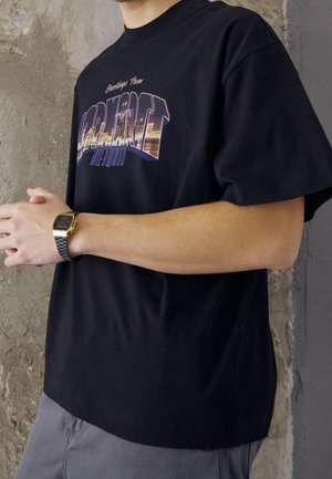 Personne portant un t-shirt noir avec l'impression du paysage urbain "Greetings from Detroit" et une montre digitale argent et or, debout contre un mur texturé.