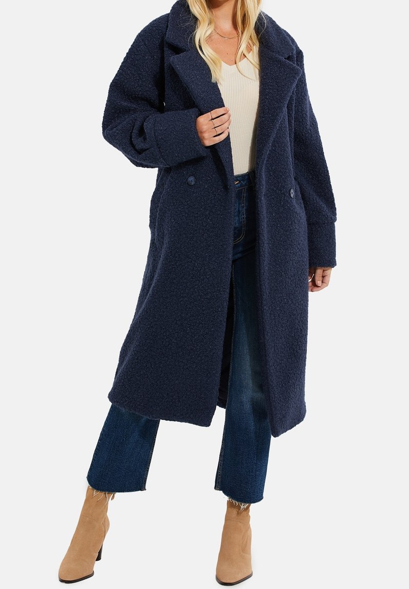 Marineblauer strukturierter Mantel mit doppelreihigem Schnitt, großen Revers und Seitentaschen, kombiniert mit ausgestellten Jeans und hellbraunen Stiefeletten.