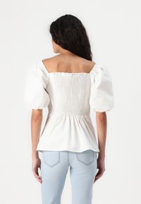 Bluză albă din bumbac, cu peplum, mâneci scurte tip puff, decolteu pătrat și detaliu smockat la spate, purtată cu blugi ajustați de culoare albastru deschis.