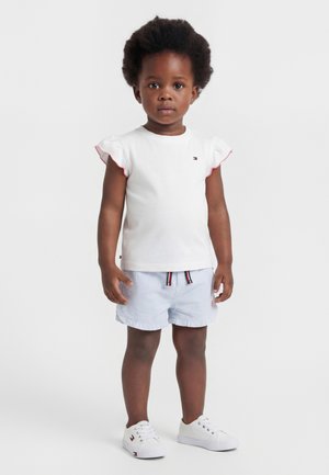 Småbarn med krøllete hår iført hvit topp med volang-ermer, blå og hvite stripete shorts og hvite joggesko på ensfarget bakgrunn.