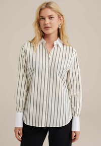 Camicia bianca con strisce verticali nere, maniche lunghe, colletto bianco e dettagli bianchi ai polsini. Presenta un orlo curvo.